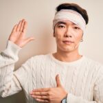 Young handsome chinese man injured for accident wearing bandage and strips on head Swearing with hand on chest and open palm, making a loyalty promise oath